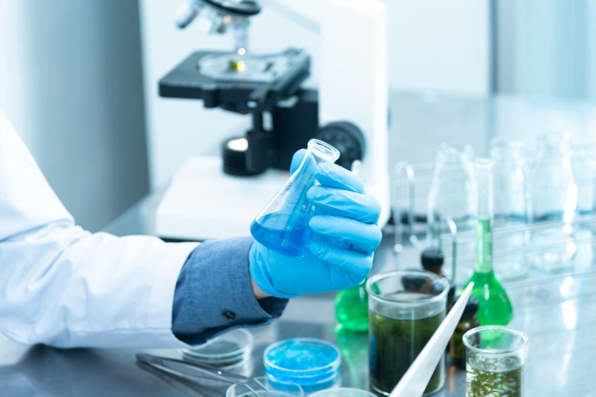 Gloved scientist inspecting a blue liquid in a flask with lab tools and microscope in background